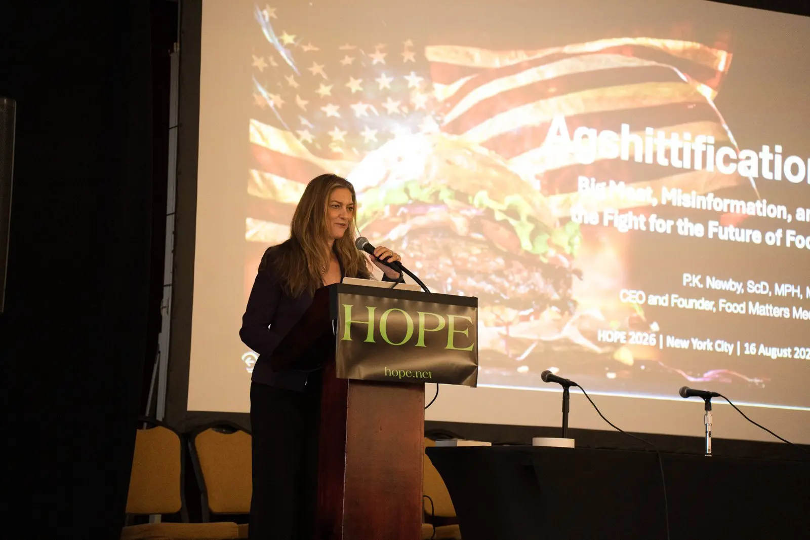 Dr. P.K. Newby speaking at the HOPE 2026 podium in front of her Agshittification title slide