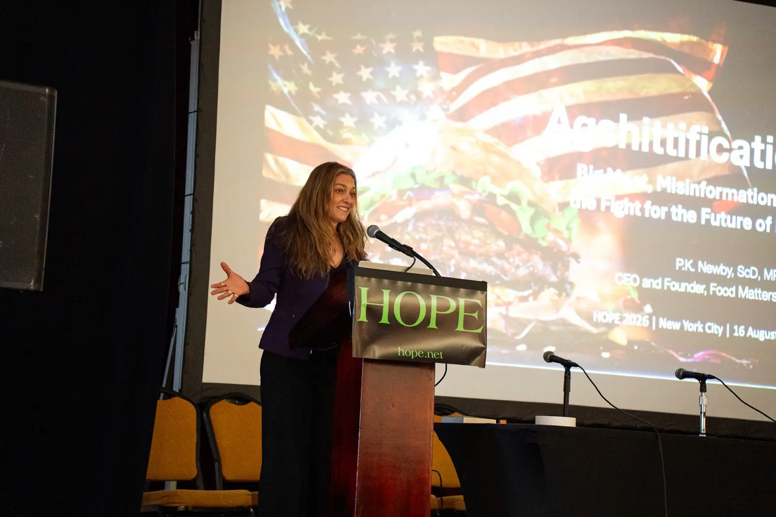 Dr. P.K. Newby gesturing at the HOPE 2026 podium in front of her Agshittification title slide
