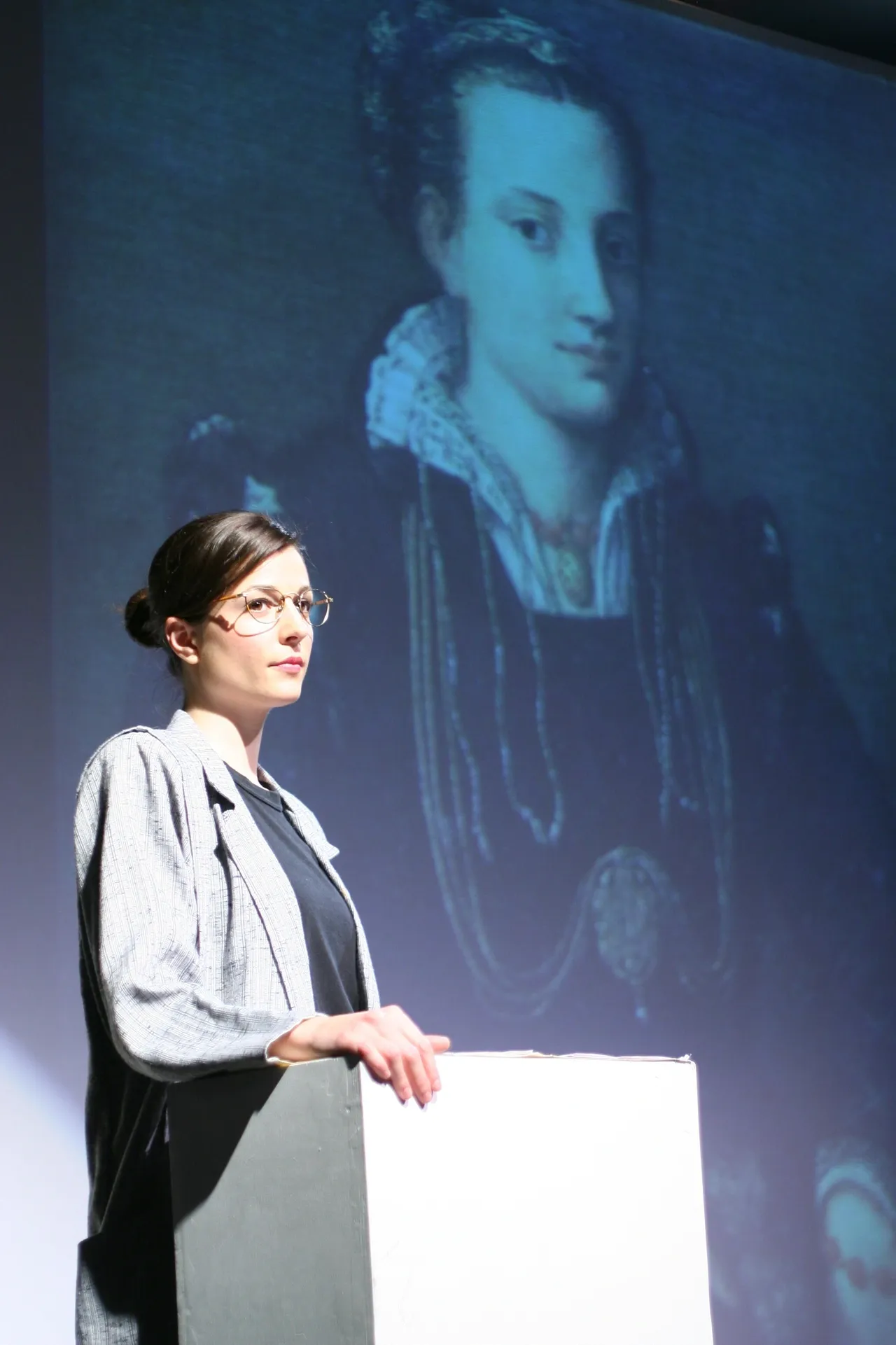 A woman gives a presentation with a historical portrait projected behind her.