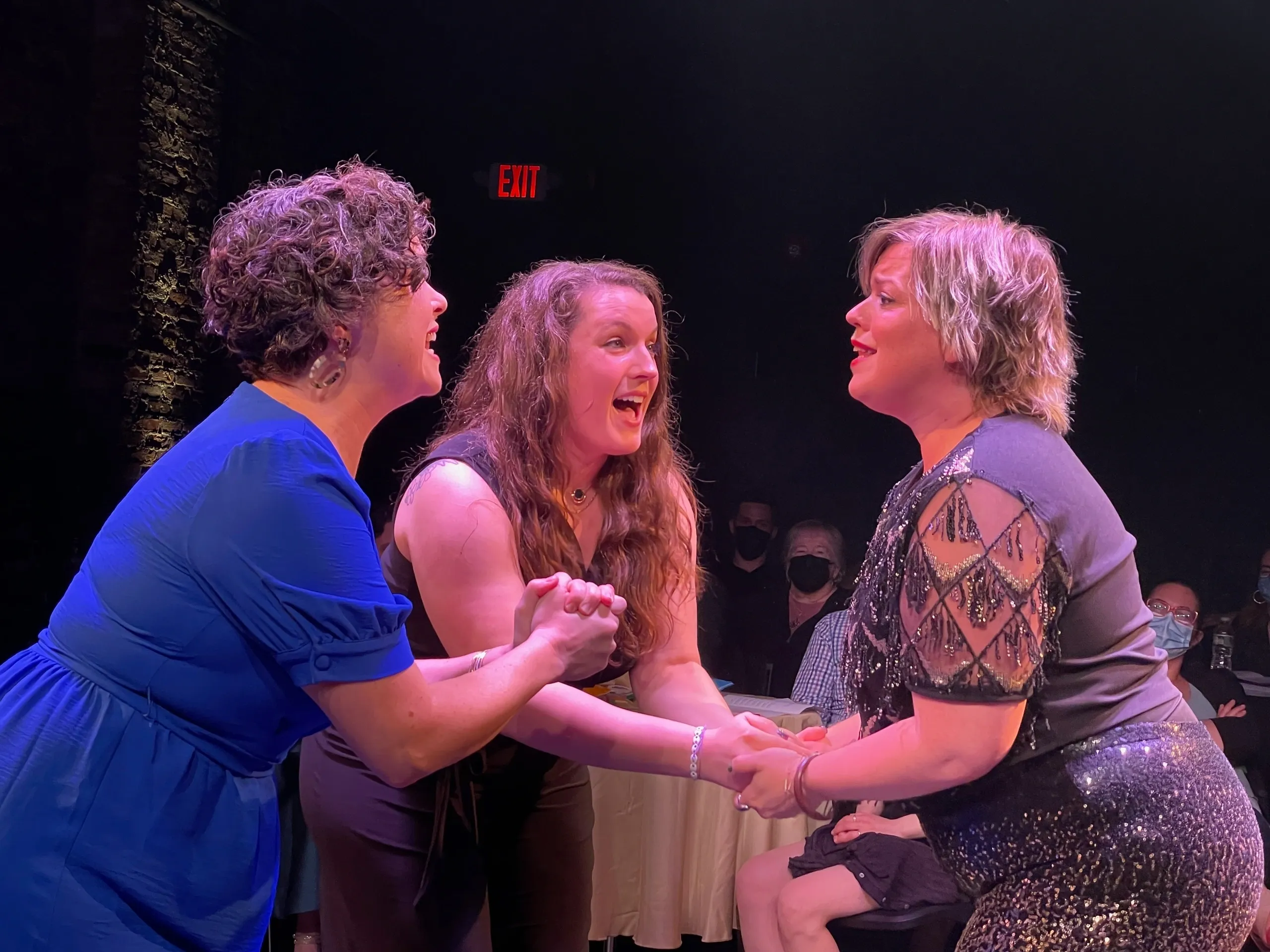 Three women holding hands and singing passionately on stage.