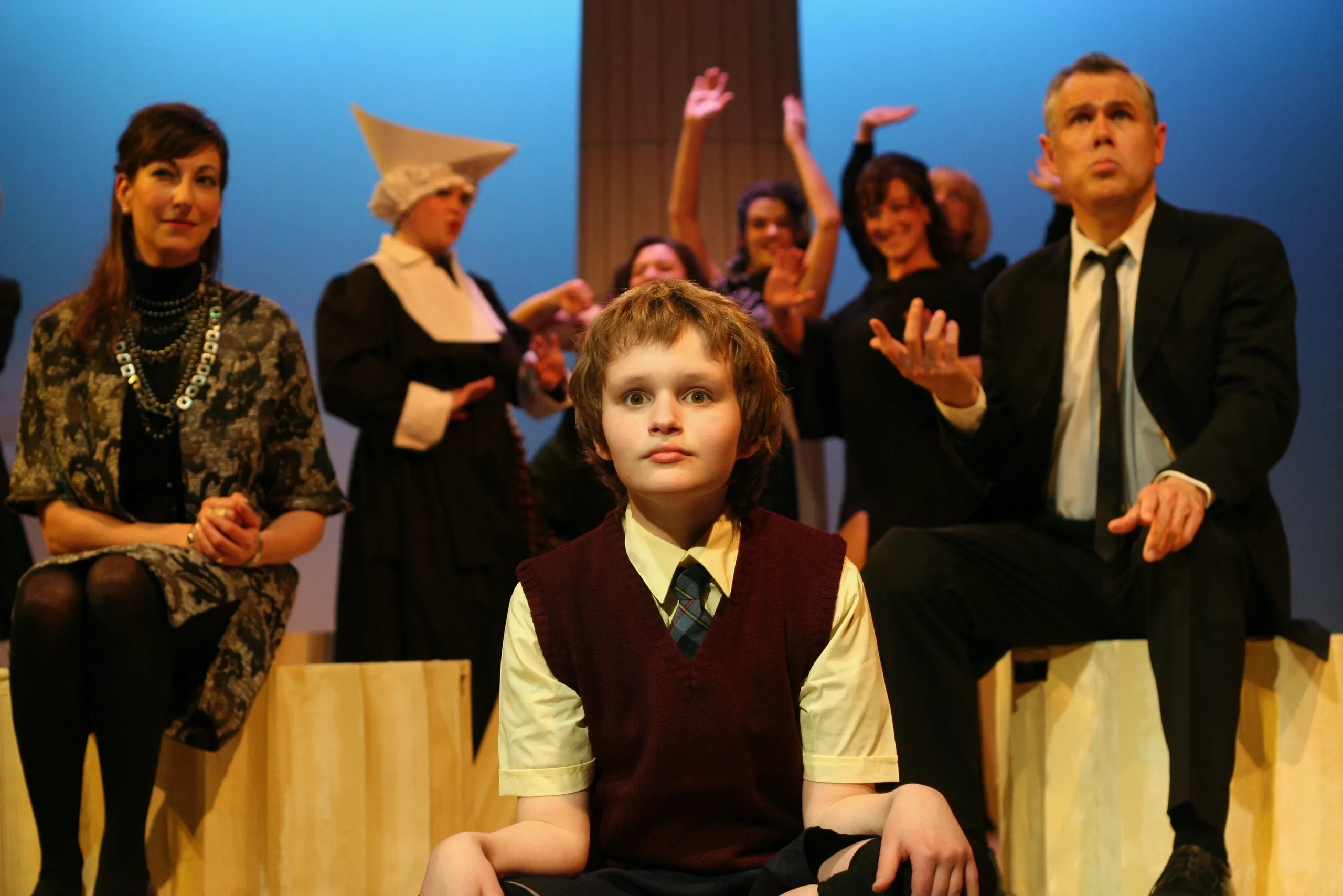 A young girl in a school uniform sits on stage with adults behind her.