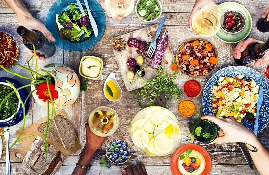 Hands sharing a colorful meal of vegetables, grains, fruit, and drinks around a dining table.