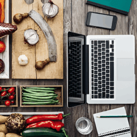 A kitchen workspace with fresh vegetables and a laptop on a wooden table.