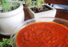 A bowl of red salsa on a table with plants in the background.