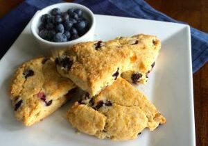 Three blueberry scones on a white plate with a small bowl of blueberries.