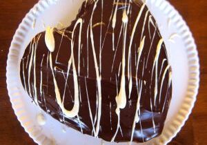 Heart-shaped chocolate cake with white drizzle on a paper plate.