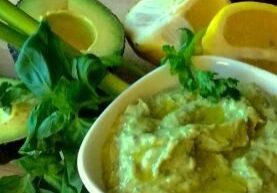 A bowl of creamy avocado dip garnished with herbs and surrounded by avocado halves and lemon slices.
