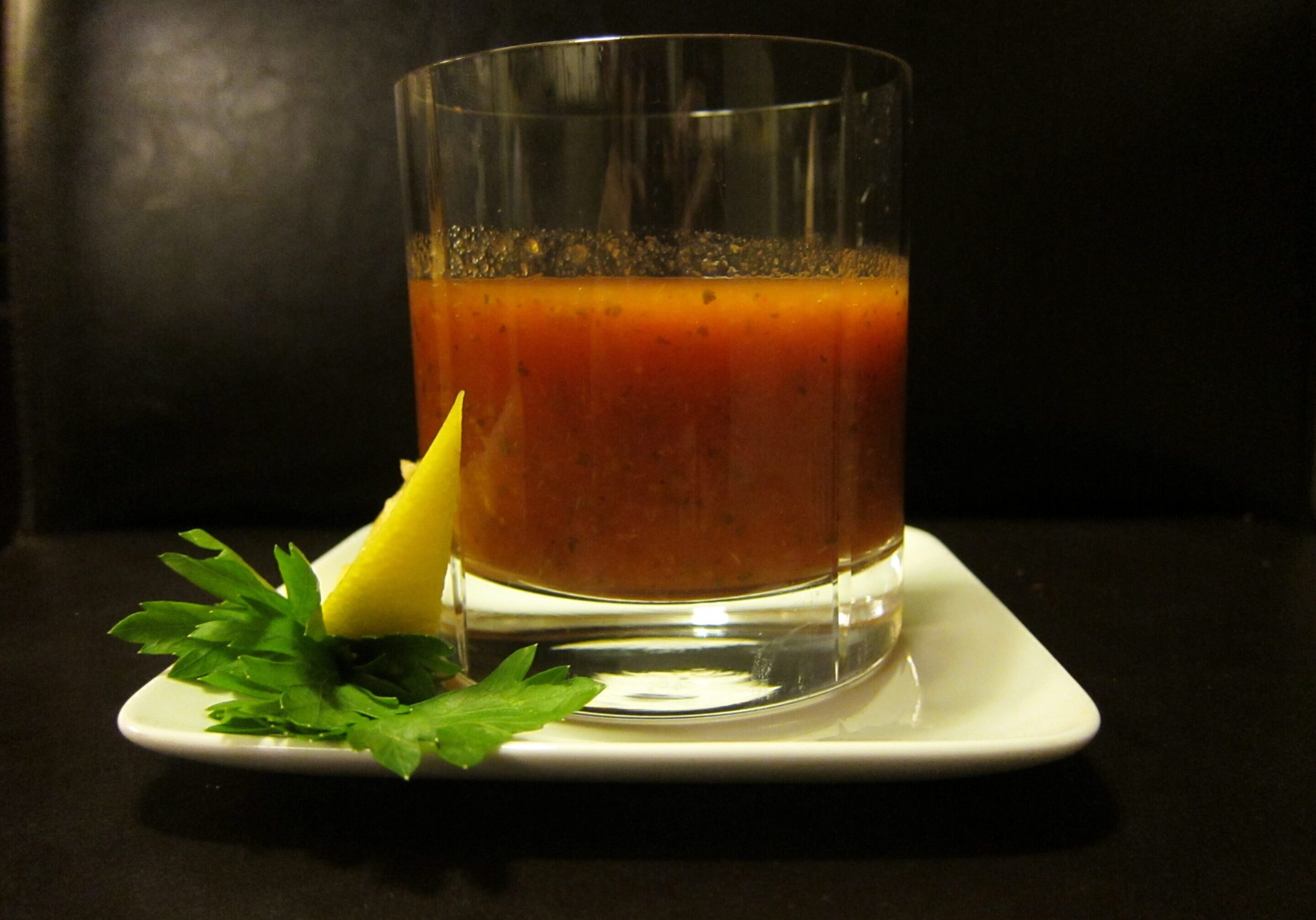 A glass of brown beverage with lemon and herbs on a white plate.