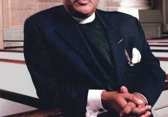 Portrait of a man in a dark suit with clasped hands.