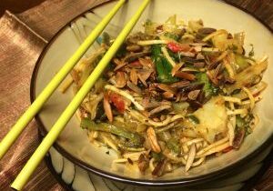A bowl of stir-fried noodles with vegetables and yellow chopsticks.
