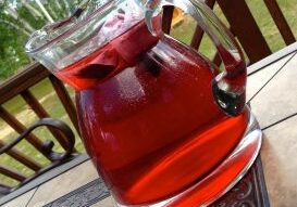 A glass pitcher filled with red fruit punch on a wooden table outdoors.