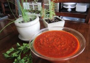 A bowl of red sauce with potted herbs on a wooden table.